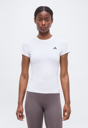 Mujer con pelo trenzado, vistiendo una camiseta ajustada blanca de Adidas y mallas grises, de pie frente a un fondo blanco liso.