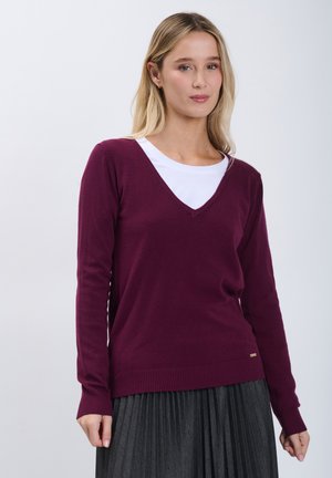 Jeune femme portant un pull col en V couleur prune par-dessus un haut blanc et une jupe plissée foncée, debout devant un fond clair uni.