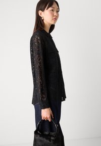 Black lace blouse with long sleeves and a collar, featuring a button-up front and floral patterns. Paired with dark denim and a small black handbag.