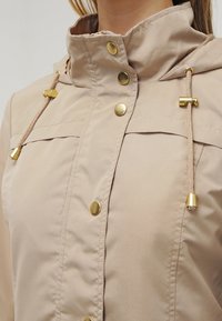 Giacca leggera beige con colletto alto, bottoni a scatto dorati e cordino decorativo. Tessuto liscio con cuciture minime e senza pattern.