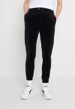 Person trägt schwarze Jogginghosen mit Kordelzug, weiße Socken und weiße Sneakers mit schwarzen Details, steht mit den Händen in den Taschen.
