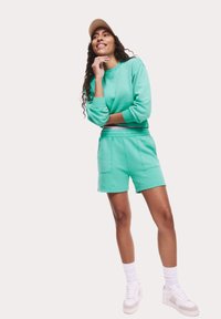 Mintgroene sweatshirt en bijpassende shorts gemaakt van zachte stof, met een relaxte pasvorm, en aangevuld met witte sneakers en een pet.