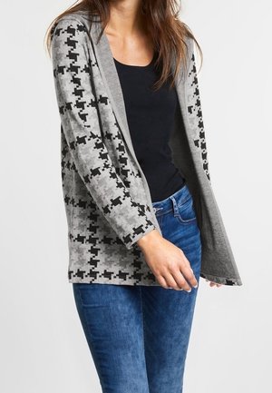 Frau trägt eine graue und schwarze Strickjacke mit Pixelmuster über einem schwarzen Oberteil und blauen Jeans, gezeigt von der Mitte der Brust bis zur Mitte des Oberschenkels.