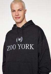 Zwarte hoodie van een katoenmix, met een voorzak, koorden en witte tekst "ZOO YORK" met een embleem erboven.