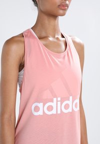 Canottiera sportiva rosa chiaro con un grande logo Adidas bianco e elementi di design triangolari, realizzata in un tessuto liscio e traspirante.