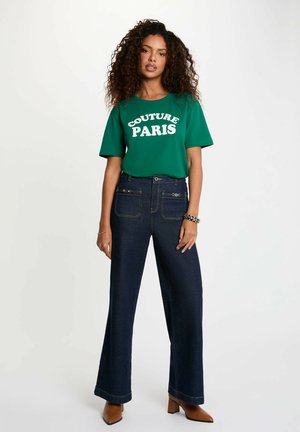 Donna con capelli ricci che indossa una maglietta verde con la scritta "Couture Paris", jeans scuri a gamba larga, scarpe con tacco marroni e braccialetti, in piedi su sfondo bianco.
