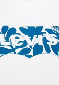 Tissu blanc avec motif floral bleu et grand texte blanc "Levi's" en gras au centre.