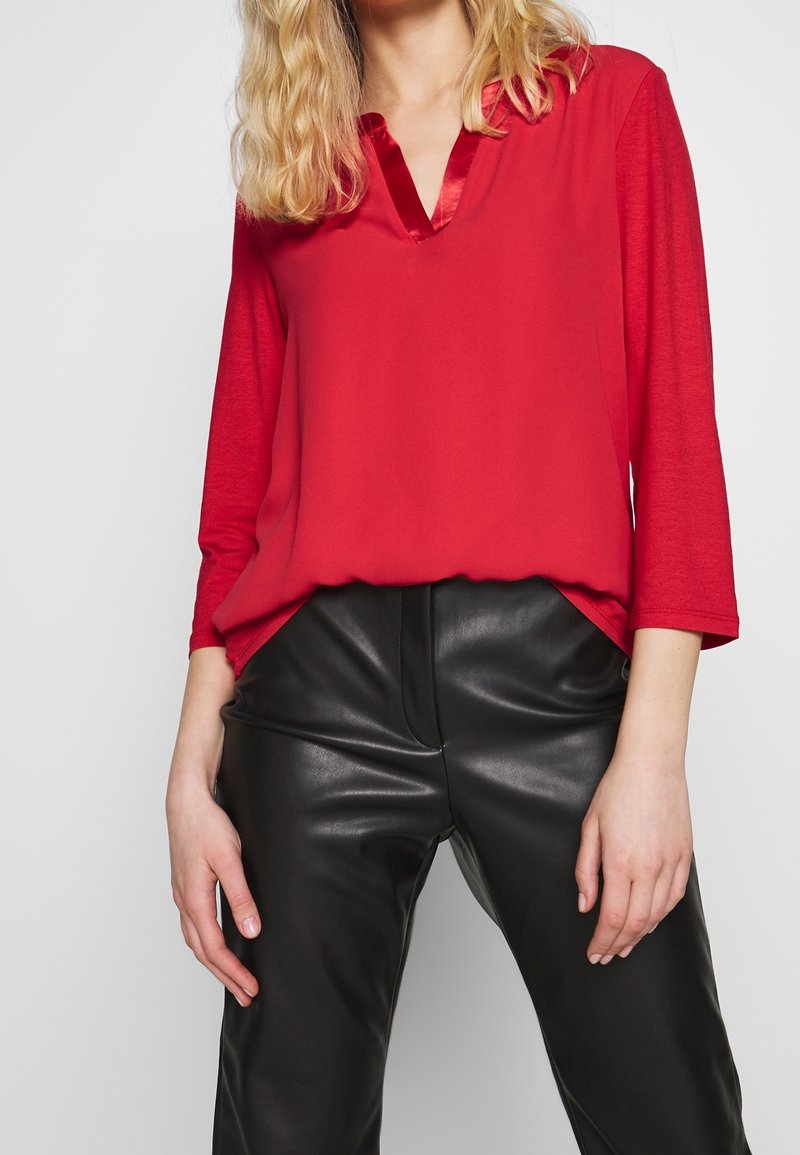 Personne blonde portant un chemisier rouge à manches longues avec un col en V en satin et un pantalon en cuir noir, debout devant un fond uni.