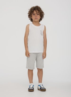 Ragazzo giovane con capelli ricci che indossa una canottiera bianca con logo "LL", pantaloncini grigi, calzini bianchi e scarpe da ginnastica nere, bianche e blu, in piedi immobile.