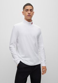 Camisa de tortuga blanca de manga larga en tela suave, con un cuello alto y un pequeño detalle de etiqueta roja en el escote, combinada con pantalones negros.