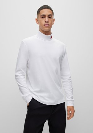 Weißes Langarm-Turtleneck-Shirt aus weichem Stoff mit hohem Kragen und einem kleinen roten Etikett am Ausschnitt, getragen mit schwarzen Hosen.