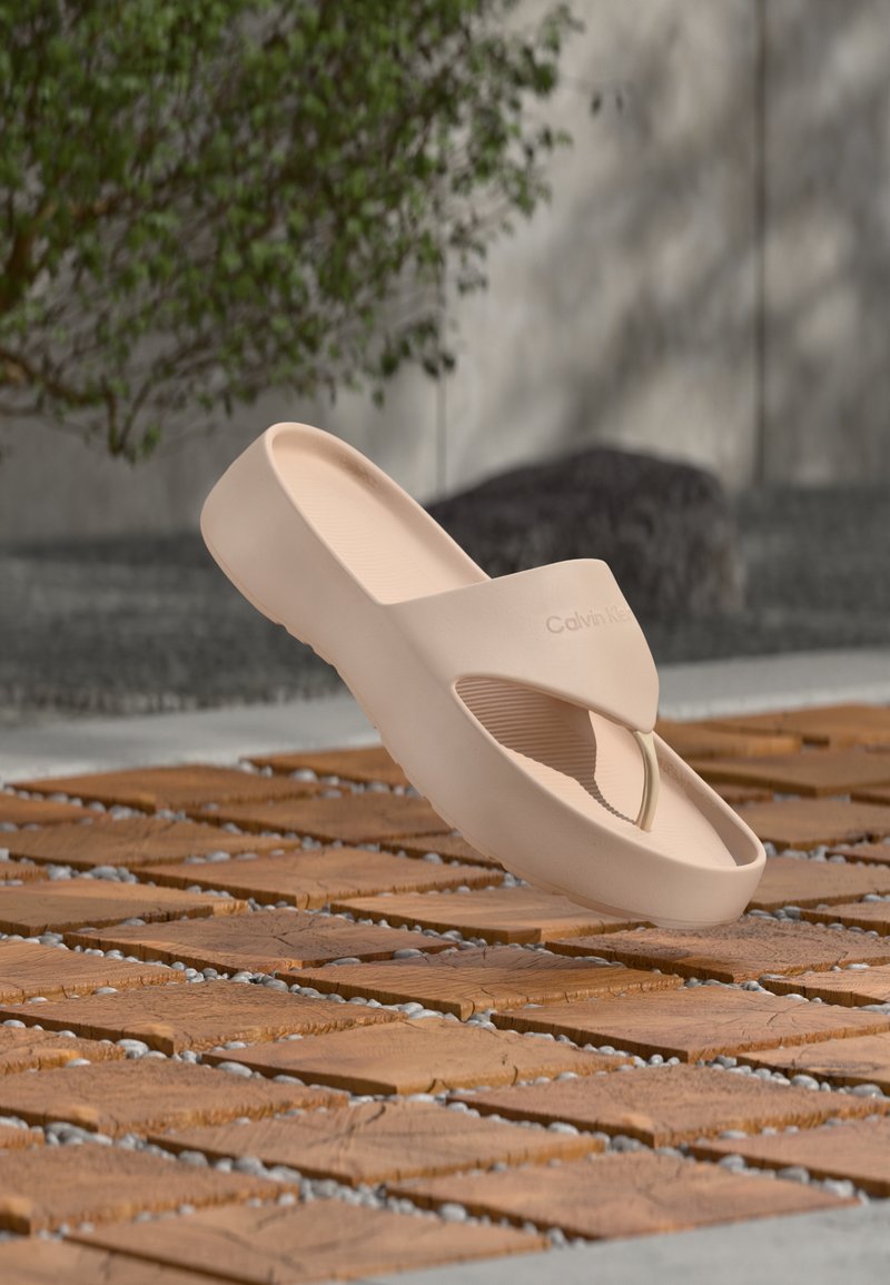 Μπεζ σανδάλι slide με επένδυση Calvin Klein που αιωρείται πάνω από ξύλινο δάπεδο με πλακάκια σε εξωτερικό χώρο, με πράσινο θάμνο και φόντο τσιμεντένινο τοίχο.