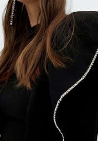 Černý oděv s texturovanou tkaninou a řaseným designem, ozdobený řadou lesklých kamínků podél okraje. Hnědé vlasy částečně viditelné.