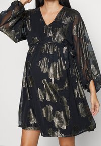 Svart klänning med abstrakt blommönster i metallisk guld, med V-ringning, genomskinliga långa ärmar och midjebälte för en luftig silhuett.