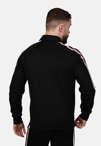 Čierna džínsová bunda na zips so zvýšeným golierom, s červenými a bielymi bočnými pruhmi. Mäkko vyrobená a priliehavá, navrhnutá pre pohodlie.