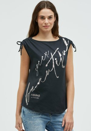 Junge Frau trägt ein schwarzes ärmelloses Oberteil mit silbernem Text und gerafften Schulterbändern, kombiniert mit hellblauen Jeans mit Textdruck.