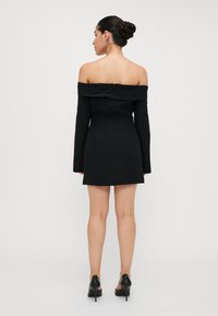 Svart av-shoulder mini-kjole med lange ermer, tettsittende kropp og glidelås bak, sammen med svarte høye hæler.