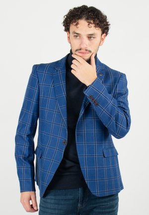 Blazer à carreaux bleu avec une coupe ajustée, revers crantés et poignets à quatre boutons. Porté sur un col roulé noir et associé à un jean.