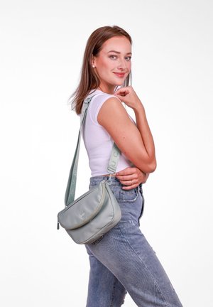 Grey crossbody bag with a soft texture, curved shape, and zip closure. Adjustable strap with logo detailing. Worn over a white top and jeans.