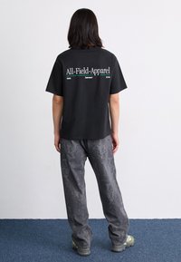 Camiseta de algodón negra con texto blanco y verde en la parte de atrás; combinada con pantalones de mezclilla grises con patrón y zapatillas gris-verde.