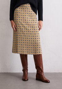 Jupe jaune avec un motif géométrique bleu, associée à des bottes en cuir marron jusqu'aux genoux, en matériau lisse, de conception mi-longue.