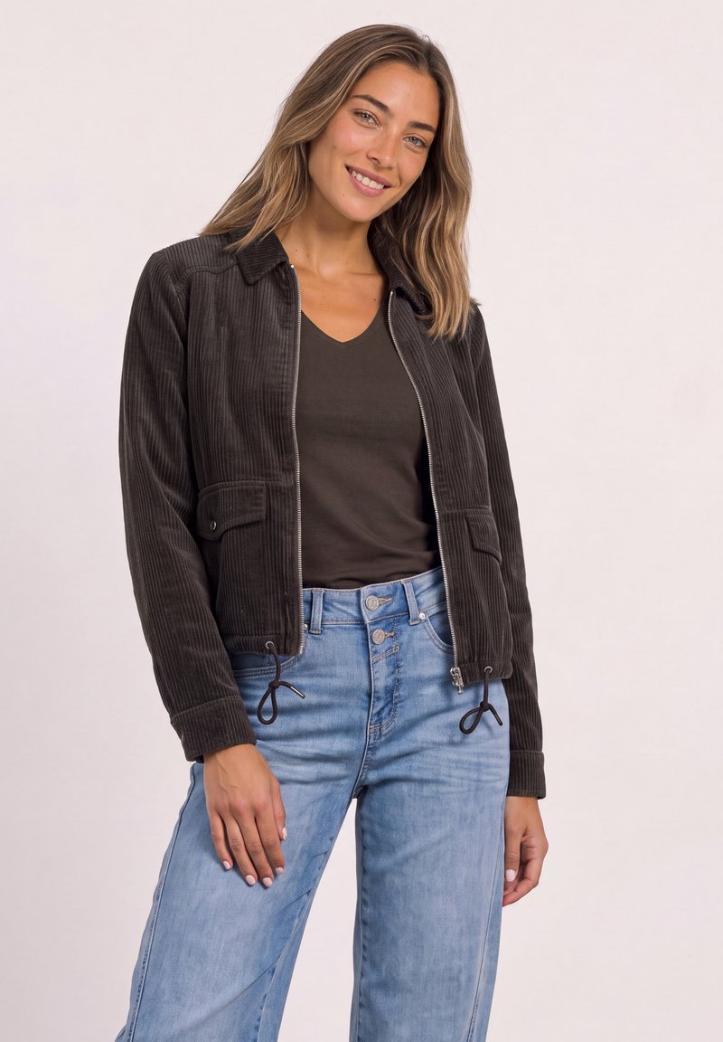 Woman smiling wearing a black corduroy zip jacket over a dark V-neck shirt and light blue high-waisted jeans against a plain background.