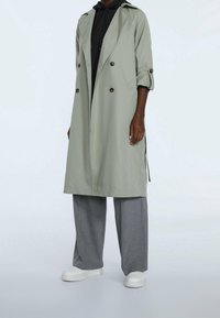 Trench coat verde claro con diseño de doble botonadura, mangas enrolladas y botones marrones, llevado sobre una sudadera negra con capucha y pantalones grises.