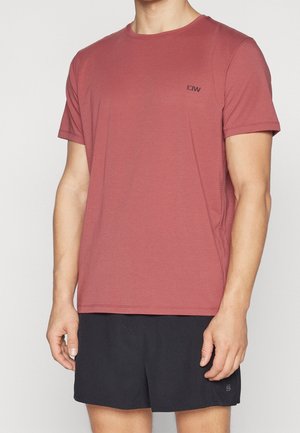 Mand iført en ensfarvet, støvet rosa T-shirt med korte ærmer og sorte sportsshorts, stående foran en hvid baggrund.