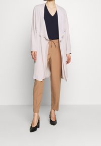 Cardigan aperto rosa chiaro con frontale drappeggiato, top blu navy a scollo a V e pantaloni beige con cordoncino. Scarpe nere con tacco completano l'outfit.