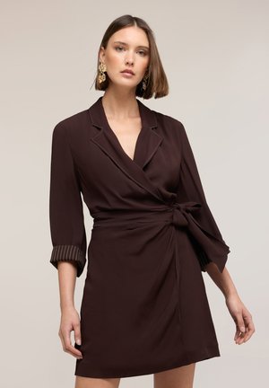 Frau trägt ein dunkelbraunes Wickelkleid mit dreiviertel Ärmeln und goldenen statement Ohrringen vor einem schlichten hellen Hintergrund.