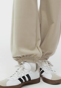 Białe trampki z czarnymi paskami i gumowymi podeszwami noszone z beżowymi joggerami i białymi skarpetami na jasnym tle.