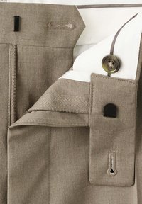Pantalon habillé marron clair avec la ceinture déboutonnée, montrant un bouton, une agrafe et une doublure intérieure blanche.