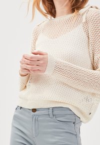 Pull en tricot crème clair, avec un tissage lâche et des poignets côtelés. Dispose de petites bretelles et d'une coupe décontractée. Assorti avec un jean bleu clair.