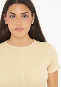 Ribbede beige top med korte ærmer, rund halsudskæring og centralt samlingsdetalje. Glat tekstur med subtile linjer og tætsiddende design.