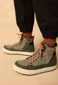 Chaussures montantes vertes avec un mélange de textures, des lacets orange, une fermeture éclair latérale et une semelle blanche épaisse. Conçues pour la durabilité et le confort.