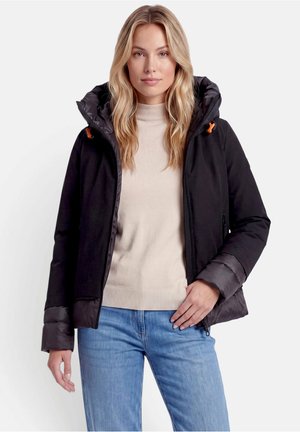 Femme blonde portant une veste noire à capuche par-dessus un pull beige et un jean bleu, debout devant un fond clair uni.
