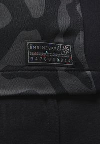 Musta vaate, jossa on sotilaspatterni, ja suorakaiteen muotoinen etiketti, jossa lukee "ENGINEERED" sekä joukko numeroita ja värikkäitä yksityiskohtia.