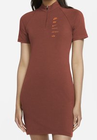 Czerwona bawełniana sukienka z wysokim kołnierzem, krótkimi rękawami i zamkiem błyskawicznym do jednej czwartej. Na froncie znajduje się pomarańczowe logo Nike oraz napisy. Gładka tekstura.