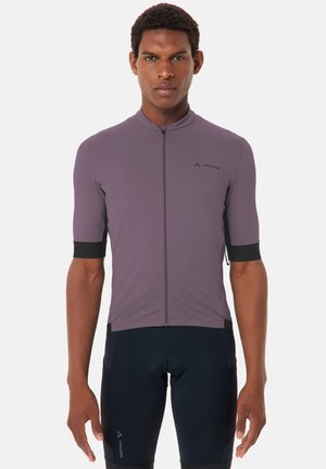 Uomo che indossa una maglia da ciclismo aderente a maniche corte di colore viola e pantaloncini da ciclismo imbottiti neri, in piedi davanti a uno sfondo bianco semplice.