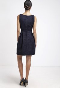 Granatowa sukienka bez rękawów z teksturowanym geometrycznym wzorem, z wcięciem w talii, plisowanym dołem i ukrytym zapięciem na plecach, w zestawie z czarnymi szpilkami.