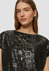 Schwarzes Oberteil mit einer glänzenden, strukturierten Oberfläche, die silberne Akzente aufweist, und einem breiten, runden Ausschnitt. Kurzärmlig mit lockerer Passform.