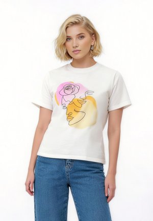 Młoda kobieta o krótkich blond włosach nosi białą koszulkę z abstrakcyjnym wizerunkiem twarzy i różą w kolorach różowym, żółtym i pomarańczowym, zestawioną z niebieskimi dżinsami.