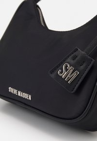 Borsa a mano nera in nylon con forma curva, dotata di un'etichetta con logo argentato e una superficie liscia. Chiusura superiore con zip e un dettaglio di branding discreto.