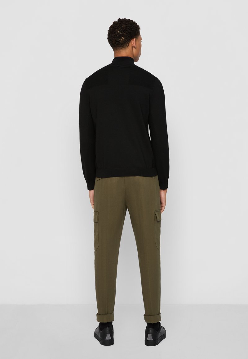 Schwarze Zip-Up-Pullover mit strukturierten Schultern, kombiniert mit olivfarbenen Cargo-Hosen mit Seitentaschen und umgeschlagenen Säumen, ergänzt durch schwarze Sneakers.