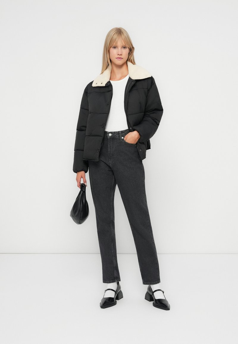 Veste rembourrée noire avec un col clair, haut blanc, jeans foncés et chaussures noires avec des accents blancs. Modèle tenant un sac à main noir.