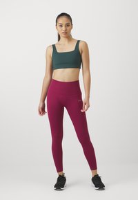 Sportsbh i mørkegrønn, kombinert med høytlivede bordeaux leggings med ribbet tekstur og slankt design. Svarte treningssko fullfører antrekket.