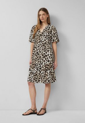 Kobieta stojąca przy jasnoszarej ścianie, ubrana w sukienkę do kolan w panterkę i czarne płaskie sandały.