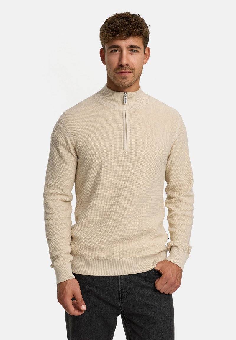 Beige zip-up sweater med høj krave, tekstureret strikmønster og ribbede ærmer, parret med mørke bukser.