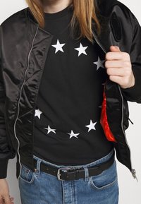 Černá saténová bomber bunda s červenou podšívkou, nosí se přes černé tričko se vzorem bílých hvězd uspořádaných do kruhového vzoru. Tmavé džíny.