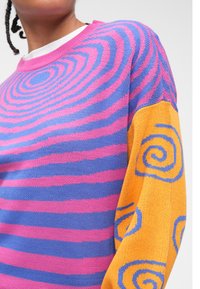 Adīta džempera, uz ķermeņa ar rozā un zilām spirālveida svītrām, un oranžas piedurknes ar zilām spirālveida dizaina elementiem. Mīksta tekstūra un brīvs piegriezums.